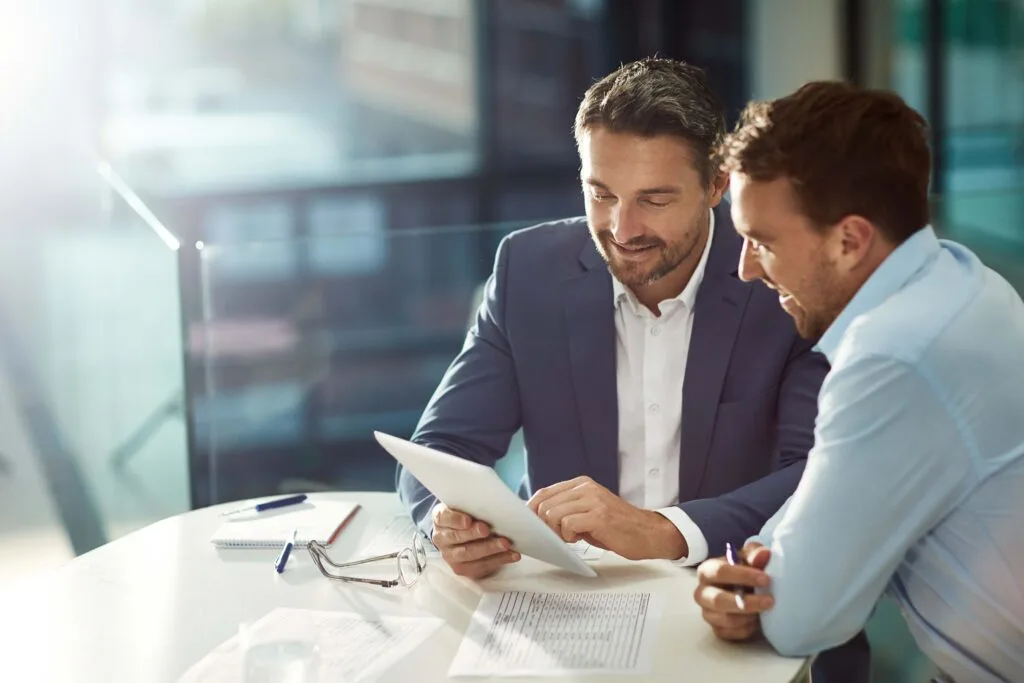 Two businessmenin a modern office discussing a tablet document at a round white table, with glasses and notebooks.